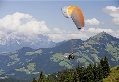 A paraglider floats over a picturesque mountain landscape. In the background, green hills and white clouds are visible.