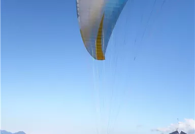 A paraglider is launching from a forested slope. In the background, mountains and a clear sky are visible.