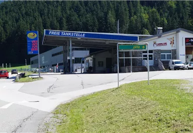 A gas station with the sign "Free Gas Station" in a green mountain landscape. In the foreground, cars and signposts can be seen.