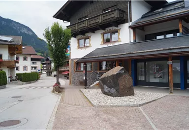 Eine ruhige Straße mit traditionellen Gebäuden und einem großen Stein in der Mitte. Im Hintergrund sind grüne Berge zu sehen.