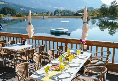 A cozy outdoor area with tables and chairs by the water. In the background, mountains can be seen under a clear blue sky.