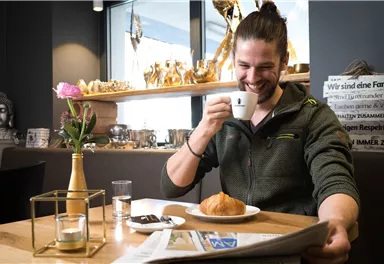 A man is sitting at a table in a café, enjoying a cup of coffee and reading a newspaper. On the table lies a croissant and a small vase with a flower.