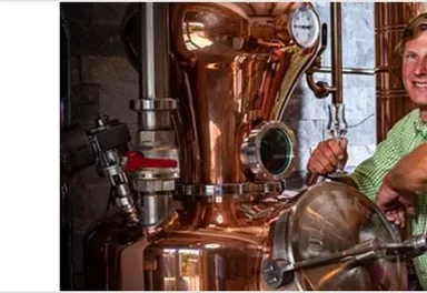 Two men are standing in front of a copper distillation pot. On the left, there is a logo with a fox.