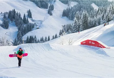 A snowboarder carries his board across snowy terrain. In the background, snow-covered mountains and trees can be seen.