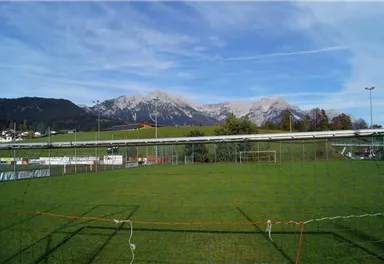 Ein Fußballfeld mit grünem Gras und einer Berglandschaft im Hintergrund. Die Berge sind teilweise mit Schnee bedeckt und der Himmel ist klar.