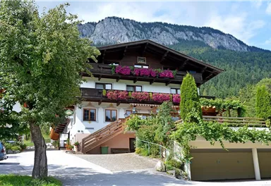 Ein charmantes Haus mit Balkonen voller Blumen. Im Hintergrund erheben sich grüne Berge unter einem klaren Himmel.