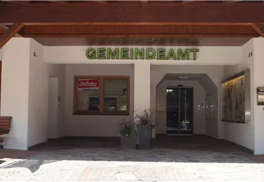 Ein Gemeindeamt mit einem einladenden Eingang. Über der Tür steht "GEMEINDEAMT" in grünen Buchstaben.