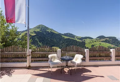 Ein malerischer Ausblick auf grüne Hügel und Berge. Auf der Terrasse stehen zwei Stühle mit Fellen und ein Tisch, ideal für eine entspannte Auszeit.
