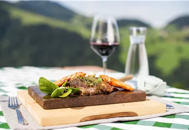 Ein delikates Gericht mit Fleisch und Garnelen auf einem Holzteller, garniert mit frischen Kräutern. Im Hintergrund ist ein Weinglas und eine schöne grüne Landschaft zu sehen.