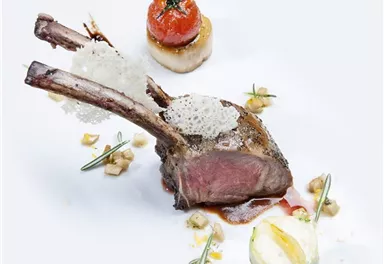 An artistically arranged dish with lamb chops, served with a stuffed tomato and vegetables. The dish is decorated with herbs and fine garnishes.