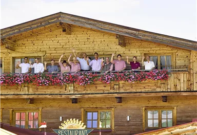 A group of people is standing on the balcony of a wooden house and waving happily. The balcony is adorned with colorful flowers.