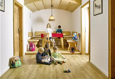 A bright room with wooden flooring, where four children are playing. At the table, another child is sitting and painting.