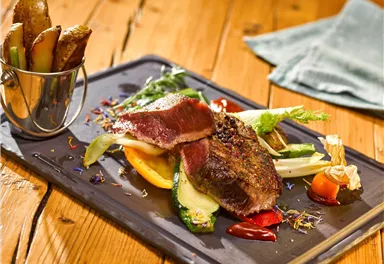 A juicy piece of meat with colorful vegetables and a side of baked potatoes. The dish is presented on a black plate and looks very appetizing.