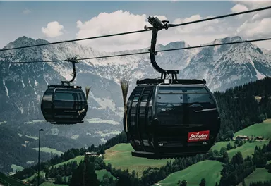 Eine Seilbahn fährt durch eine malerische Berglandschaft. Im Hintergrund sind hohe Berge und grüne Wiesen zu sehen.