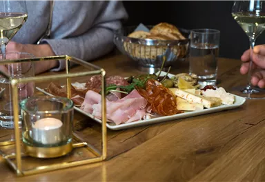 A table with a delicious snack platter that includes various types of cheese, ham, and olives. In the background, there are glasses of wine and water.