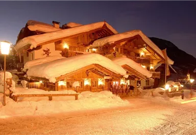 A traditional chalet in the snow with a snow-covered roof. The warm lighting creates a cozy atmosphere in the wintry landscape.