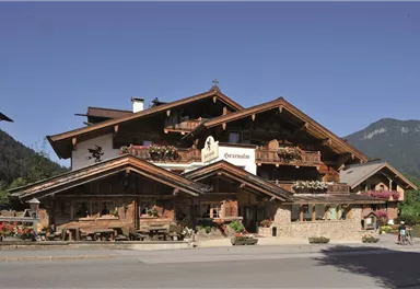 Ein traditionelles Alpenhaus mit Holzfassade und Balkonen. Im Vordergrund befindet sich eine Terrasse mit Sitzgelegenheiten und blühenden Blumen.