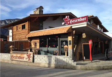 A rustic bar called "Hexenbar" with an inviting outdoor area. The facade is made of wood and stone, surrounded by mountains.