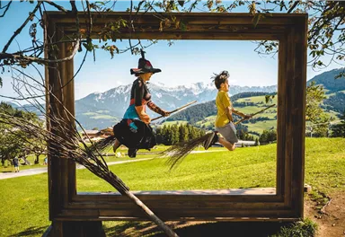 Zwei Kinder fliegen auf Besen durch einen Holzrahmen inmitten einer schönen Landschaft. Im Hintergrund sind Berge und ein klarer Himmel sichtbar.