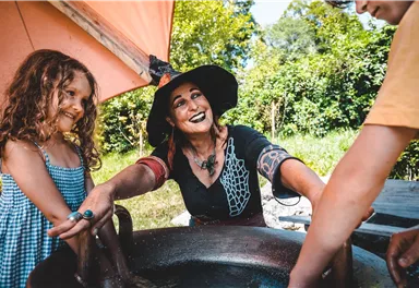 Eine freundliche Frau mit einem schwarzen Hut lacht, während sie mit zwei Kindern in einem großen Behälter spielt. Im Hintergrund sind Bäume und grünes Gras zu sehen.