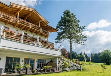A beautiful chalet with a wooden balcony and a view of the green landscape. In the foreground, there are plants and a stone wall.