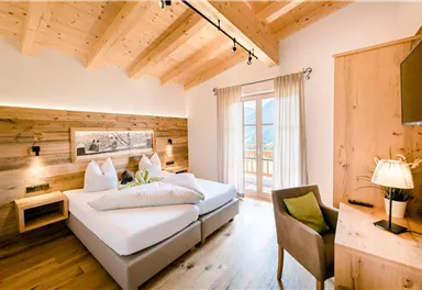 A cozy hotel room with wooden walls and large windows. There are two single beds, a desk, and a modern television.