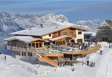 A cozy mountain hut in the snow, surrounded by impressive mountains. Many people enjoy the winter sun on the terrace.