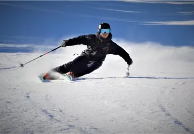 Ein Skifahrer macht eine Kurve auf frischem Schnee. Im Hintergrund sind schneebedeckte Berge und ein klarer Himmel zu sehen.