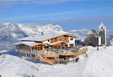 A snow-covered mountain village with a modern restaurant and an old church. The surroundings are surrounded by majestic mountains and a clear sky.