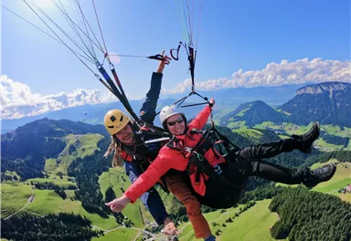 Two people paragliding over a green, hilly landscape. The sky is blue with some clouds.