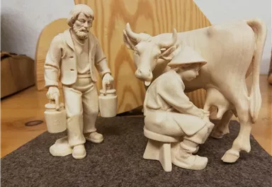 Eine geschnitzte Holzfigur zeigt einen Bauern mit zwei Eimern neben einem Mädchen, das eine Kuh melkt. Die Szene vermittelt ländliches Leben und Tradition.