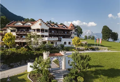 A picturesque hotel in alpine style, surrounded by green meadows and trees. Majestic mountains can be seen in the background.