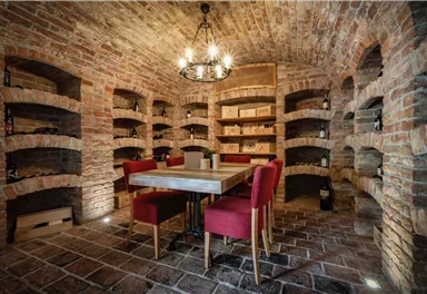 Ein stilvoller Weinkeller mit Backsteinwänden und Regalen für Weinflaschen. In der Mitte steht ein Tisch mit roten Stühlen, ideal für Weinproben.