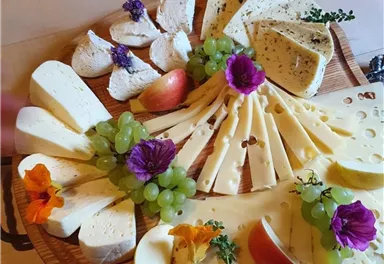Eine kunstvoll angelegte Käseplatte mit verschiedenen Käsesorten, Trauben und bunten Blumen. Die Platte ist auf einem Holzbrett arrangiert und bietet eine ansprechende Präsentation.
