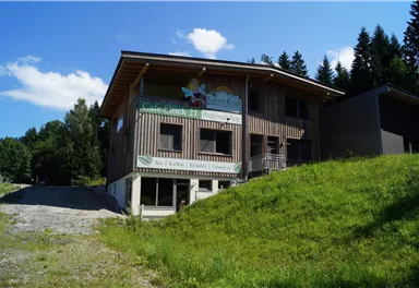 A modern wooden building in a natural environment. In the background, there are trees and a blue sky.