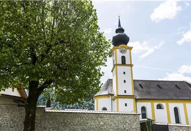 A church with a distinctive tower stands next to a large tree. The sky is partly cloudy and the buildings are surrounded by a stone wall.