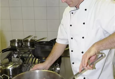 Ein Koch bereitet ein Gericht in einer professionellen Küche zu. Er rührt in einem großen Topf auf dem Herd.