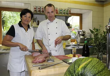 Ein Koch und eine Köchin arbeiten in einer modernen Küche. Sie schneiden Fleisch und bereiten Zutaten für ein Gericht vor.