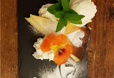 An artistically arranged dessert on a black plate. It consists of ice cream, cream, fruits, and edible flowers.