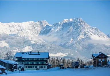 A picturesque winter landscape with snow-covered mountains and a bright blue sky. In the foreground are two houses, also standing in the snow.