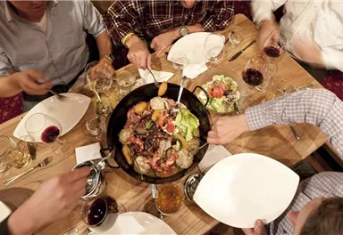 A cheerful dining gathering with several people gathered around a large plate with various dishes. There are glasses with drinks and empty plates visible on the table.