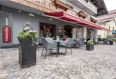 A cozy outdoor area of a café with tables and chairs. Red umbrellas provide shade, and decorative plants are also present.