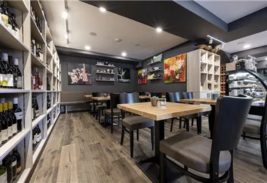 A modern restaurant with wooden tables and black chairs. Shelves with wine bottles and colorful artworks decorate the walls.