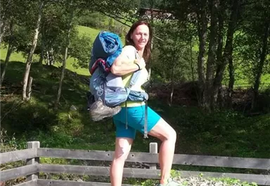 A woman is standing on a small hill with a backpack. In the background, there are trees and a green landscape.