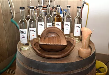A wooden bowl and a wooden pitcher are placed on a wine barrel, around which bottles with clear liquids are arranged. The scene conveys a rustic and handcrafted atmosphere.