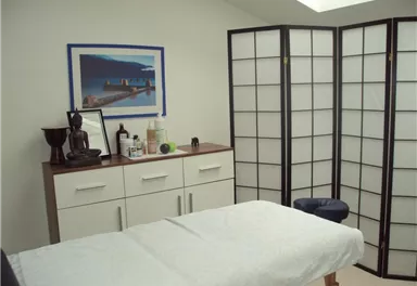 Ein ruhiger Raum mit einem Behandlungstisch und einem Paravent. An der Wand hängen ein Bild und einige Utensilien, die Entspannung fördern.