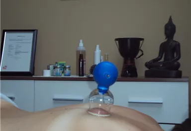 Eine therapeutische Behandlung mit einer Schröpfkopfmethode auf dem Rücken einer Person. Im Hintergrund sind verschiedene Produkte und eine Buddha-Statue zu sehen.