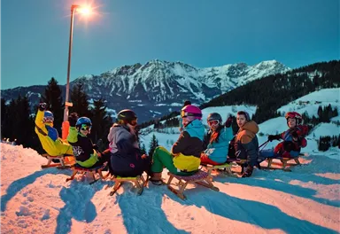 Eine Gruppe von sieben Personen sitzt auf Schlitten im Schnee. Im Hintergrund sind Berge und ein klarer Abendhimmel zu sehen.