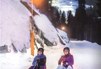 Zwei Personen sitzen auf einem Schlitten in der verschneiten Landschaft. Es ist Abend und die Umgebung ist von Lichtern beleuchtet.
