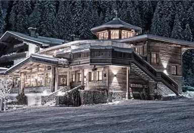 A cozy wooden house in the snow, surrounded by tall fir trees. The surroundings are wintry and peaceful.